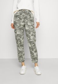 Spodnie cargo w kamuflażowym wzorze w odcieniach zieleni i szarości. Posiadają elastyczne mankiety, boczne kieszenie oraz zapięcie na guzik. Noszone z białymi trampkami.