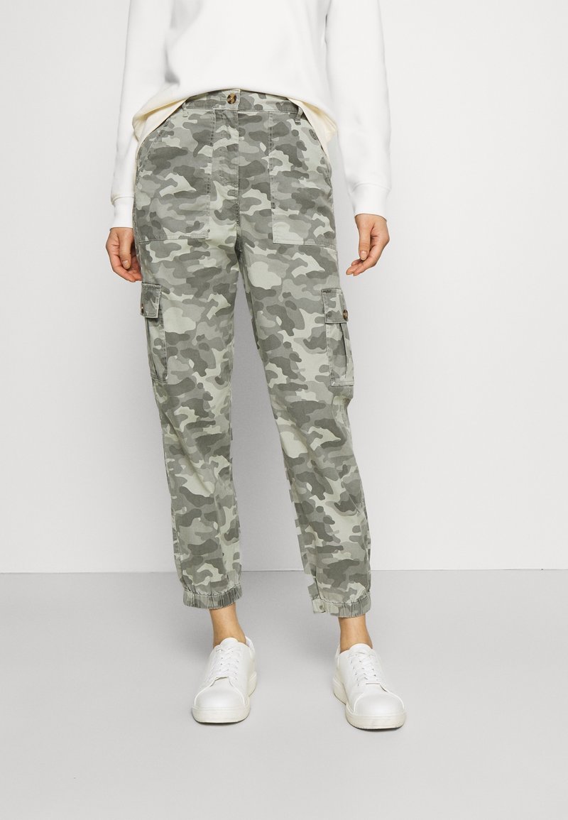 Spodnie cargo w kamuflażowym wzorze w odcieniach zieleni i szarości. Posiadają elastyczne mankiety, boczne kieszenie oraz zapięcie na guzik. Noszone z białymi trampkami.