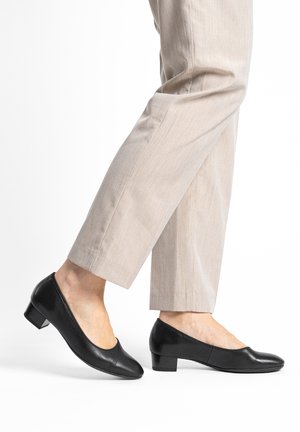 Black leather low-heeled shoes with a rounded toe and a smooth finish, worn with light-colored, tailored cropped pants.