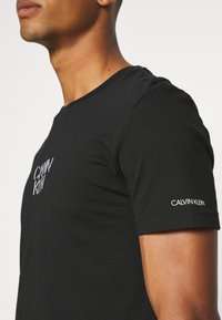 Camiseta de algodón negra con cuello redondo. Presenta el logo blanco "CALVIN KLEIN" en el pecho y la manga. Mangas cortas y textura suave.
