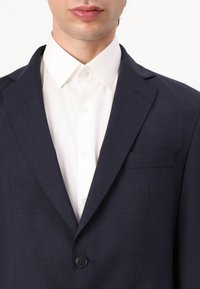 Blazer azul marino hecho de tejido texturizado, con un botón y un bolsillo en el pecho. Debajo, se puede ver una camisa blanca con cuello.