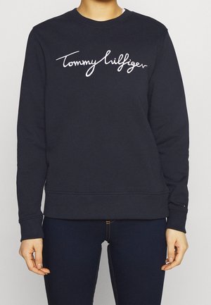 Osoba ubrana w czarną bluzę Tommy Hilfiger z białym kursywnym logo i ciemne spodnie, stojąca na jasnym jednolitym tle.
