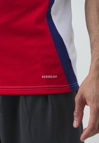 Cămașă sport roșie cu accente bleumarin și alb, având o textură din plasă pe lateral și branding "AEROREADY" aproape de tiv.