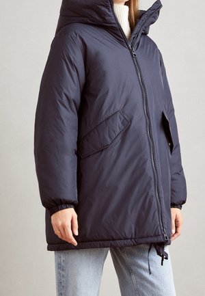 Kobieta ubrana w granatową puchową kurtkę z przednim suwakiem, bocznymi kieszeniami z patką, elastycznymi mankietami i wysokim kołnierzem, nosząca pod nią biały sweter i jasne dżinsy.