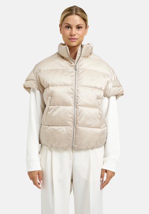Frau trägt eine beige gesteppte Kurzarmjacke über einem weißen Langarmshirt und weißen Hosen, steht vor einem schlichten Hintergrund.