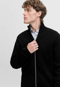 Sort zip-up sweater lavet af struktureret stof, med høj krave og en sølvfarvet lynlås, båret over en blå og hvid stribet skjorte.