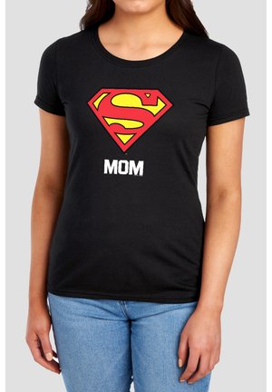 Woman wearing black t-shirt with red and yellow Superman emblem and white text "MOM," paired with light blue jeans against a plain background.