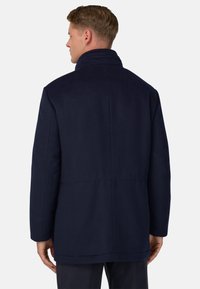 Un homme portant un manteau bleu marine foncé est debout, le dos à la caméra, devant un fond clair et uni.