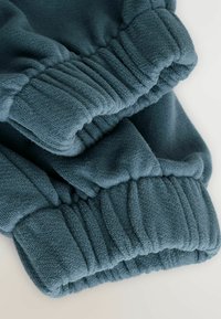 Primo piano di pantaloni tuta in felpa di un verde teal scuro con polsini elasticizzati e tessuto testurizzato su uno sfondo chiaro.