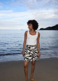Femme aux cheveux bouclés portant un débardeur blanc et une jupe à motifs noir et blanc, debout sur une plage près d’une eau océanique calme.