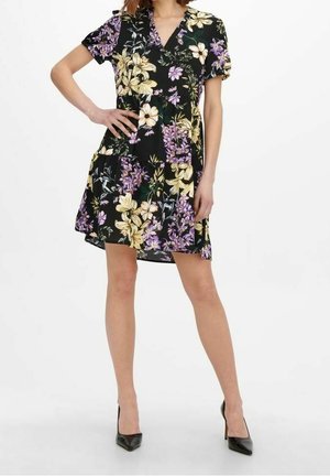 Frau trägt ein schwarzes kurzärmeliges Blumenmusterkleid mit gelben und violetten Blumen und schwarzen High Heels, steht mit einer Hand in der Hüfte.