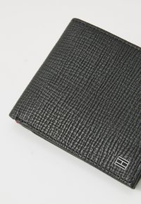 Portefeuille en cuir noir avec une surface texturée, arborant un accent logo argenté. Forme rectangulaire compacte avec des bords cousus.