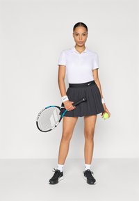 Λευκό πόλο μπλούζα και μαύρη πλισέ φούστα τένις, κρατώντας ρακέτα και μπάλα τένις, με μαύρα αθλητικά παπούτσια και βραχιόλια καρπού.