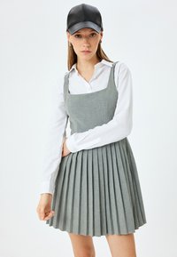 Robe plissée grise avec un décolleté carré, portée sur une chemise blanche. Le mannequin porte une casquette en cuir noir. Texture lisse et design ajusté.
