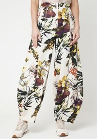 Brede broek met kleurrijke bloemen- en bladerenpatronen, gecombineerd met beige sneakers, gedragen door een staande persoon tegen een effen achtergrond.