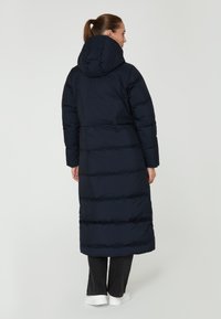 Abrigo de plumas largo en color navy con capucha, que presenta un diseño acolchado y puños elásticos. Llevado sobre pantalones negros y zapatillas blancas.