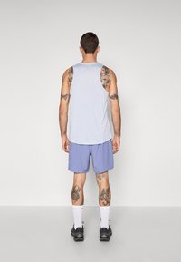 Débardeur gris clair et shorts violet clair, avec une coupe décontractée. Chaussettes blanches et chaussures de sport noires complètent la tenue. Tatouages visibles sur les bras et les jambes.