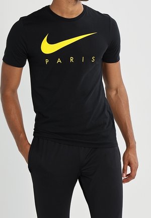 Mann trägt schwarzes Nike-T-Shirt mit gelbem Swoosh und dem Schriftzug "PARIS", kombiniert mit schwarzen Hosen, steht mit einer Hand in der Tasche.