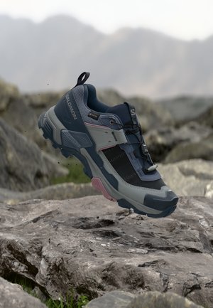 Chaussure de randonnée Salomon bleue et grise avec un mesh noir et des touches roses sur un terrain rocailleux, en l'air comme en train de marcher ou de sauter.
