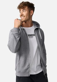 Sweat à capuche gris zippé avec une capuche, des poignets côtelés et une fermeture éclair blanche. Porté sur un t-shirt blanc avec un logo noir. Texture de tissu lisse.