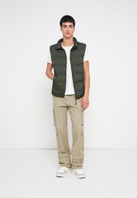 Zelená prešívaná vesta s vysokým golierom, nosená cez biele tričko. Kombinovaná s béžovými cargo nohavicami a bielymi teniskami. Jednoduchý dizajn bez vzorov.