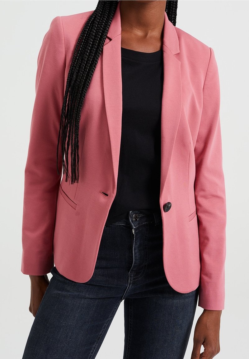 Růžový blazer s konfekčním střihem, hladkou tkaninou, jedním knoflíkem a klopami, kombinovaný s černou košilí a tmavými džíny.