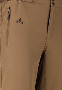 Ruskeat ulkohousut, joissa vasemmassa reisitaskussa vetoketjullinen tasku ja pienen mustan kolmionmuotoinen logo taskun yläpuolella.