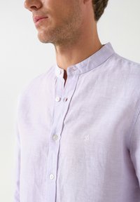 Camisa de lino lavanda con cuello alzado, que cuenta con una tapeta de botones y un logo bordado en el pecho. Textura suave y ligero peso.