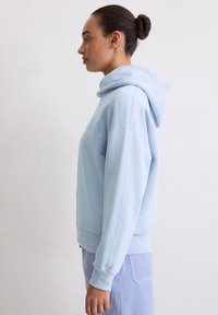 Kobieta z ciemnymi włosami upiętymi w kok, ubrana w jasnoniebieską bluzę z kapturem i niebieskie spodnie sztruksowe, stojąca bokiem na tle gładkiej, białej ściany.