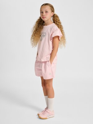Junges Mädchen mit langen lockigen Haaren steht und trägt ein hellrosa T-Shirt, passende Shorts, weiße knielange Socken und rosa Turnschuhe auf grauem Hintergrund.