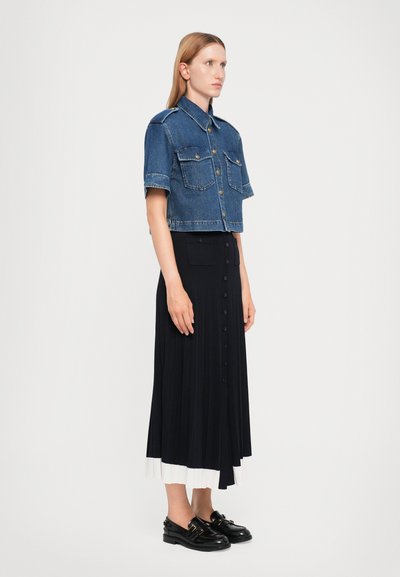 Kuttet denimjakke med to brystlommer, kombinert med en svart plisserte skjørt med hvit panel ved kanten og svarte slip-on sko.