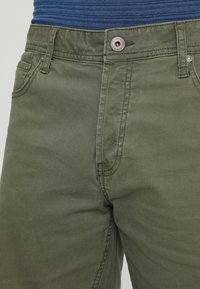 Nahaufnahme von olivgrünen Hosen mit Knopf- und Reißverschluss, sichtbaren Gürtelschlaufen, getragen mit einem blauen gerippten Shirt, das hinein gesteckt ist.