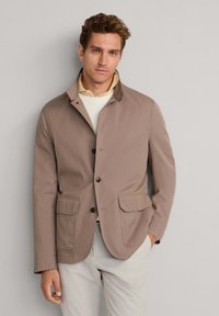 Veste marron clair à boutons avec col, deux poches frontales et texture de tissu lisse. Portée sur un pull crème.