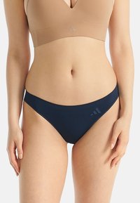 Frau in einem beigen Sport-BH und navyblauen Bikini-Unterhosen mit dezentem Logo vor einem einfarbigen Hintergrund.