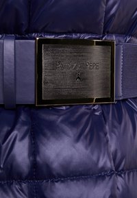 Navy dunjakke sektion med quiltet tekstur, der har et bredt stofbælte og en rektangulær metallisk logo-plade med indgraveret mærkenavn.