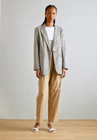 Karierter Blazer mit lockerer Passform, beigen Hosen und weißen Sandalen. Der Stoff ist strukturiert mit einer Mischung aus Farben im Muster.