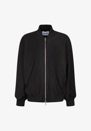 Homme portant une veste bomber noire zippée et un pantalon clair, debout devant un fond uni.