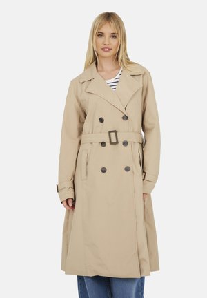 Femme portant un trench-coat beige à double boutonnage avec ceinture et boutons noirs, debout devant un fond clair uni.