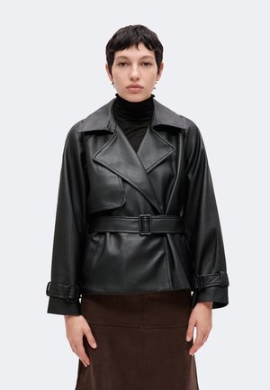 Personne aux cheveux courts et foncés portant une veste en cuir noire ceinturée et une jupe marron, debout devant un fond clair uni.