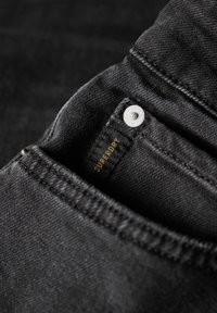 Los vaqueros de mezclilla negros presentan un bolsillo delantero con un botón plateado y una etiqueta que dice "SUPERDRY" en dorado. La tela tiene una textura suave.