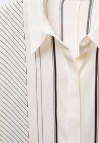 Chemise blanche à col avec des rayures noires verticales et des fines lignes noires diagonales sur le côté gauche, boutonnée près du col.