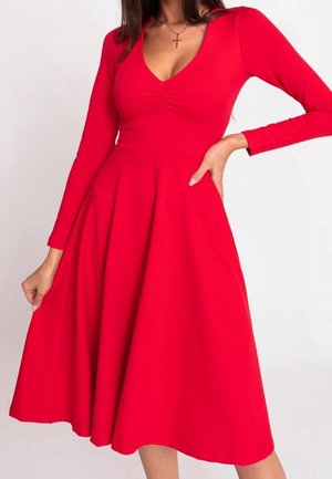 Frau trägt ein langärmliges rotes A-Linien-Kleid mit V-Ausschnitt und gerafftem Detail an der Brust, posiert mit einer Hand in der Hüfte und der anderen, die den Rock hält.