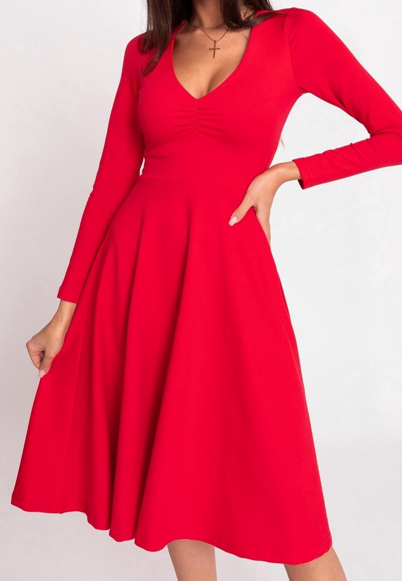 Femme portant une robe rouge à manches longues, coupe trapèze, avec un col en V et un détail froncé au niveau de la poitrine, posant avec une main sur la hanche et l’autre tenant la jupe.