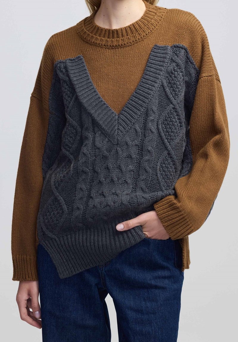 Bruine en grijze gebreide trui met een gestructureerd kabelpatroon, voorzien van een diepe V-hals en geribbelde manchetten, gecombineerd met blauwe denim.