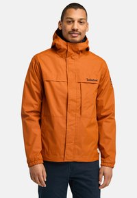 Orange vattentät jacka med hög krage, justerbar huva, två framfickor och elastiska ärmslut, med Timberland-logotyp på bröstet.