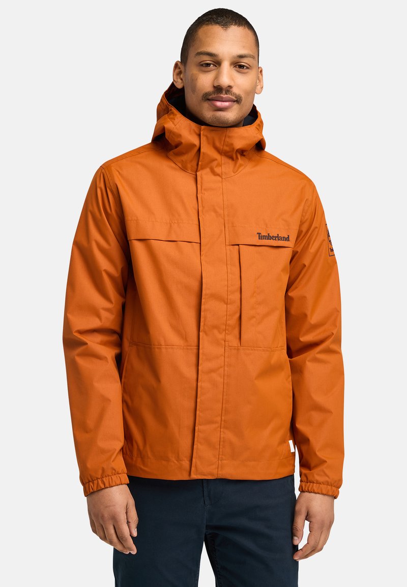 Orange vattentät jacka med hög krage, justerbar huva, två framfickor och elastiska ärmslut, med Timberland-logotyp på bröstet.