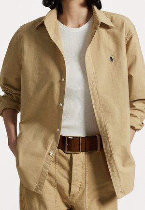 Personne portant une chemise beige boutonnée par-dessus un débardeur blanc côtelé avec un pantalon beige et une ceinture marron, mains dans les poches, manches de la chemise retroussées.