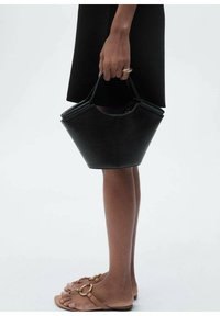 Black leather handbag with a geometric silhouette, short handles, and smooth texture, held at the side beside a short black dress and sandals.