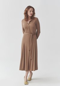 Langes braunes Kleid mit Knopfleiste, Bindegürtel und gefüttertem Rock. Weicher Stoff und lockerer Schnitt. Kombiniert mit hellen Schuhen.