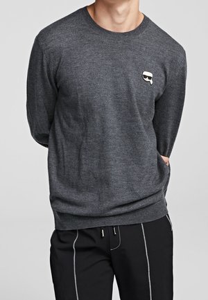 Mand iført en mørkegrå sweater med et lille patch med et ansigt med solbriller på brystet, kombineret med sorte bukser med hvide syninger.